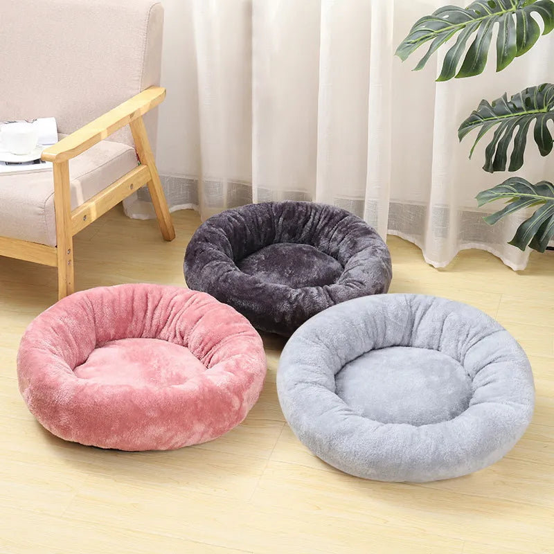 Three plush round pet beds in pink, dark gray, and light gray arranged on a light wood floor in a bright room with curtains, a chair, and a plant.