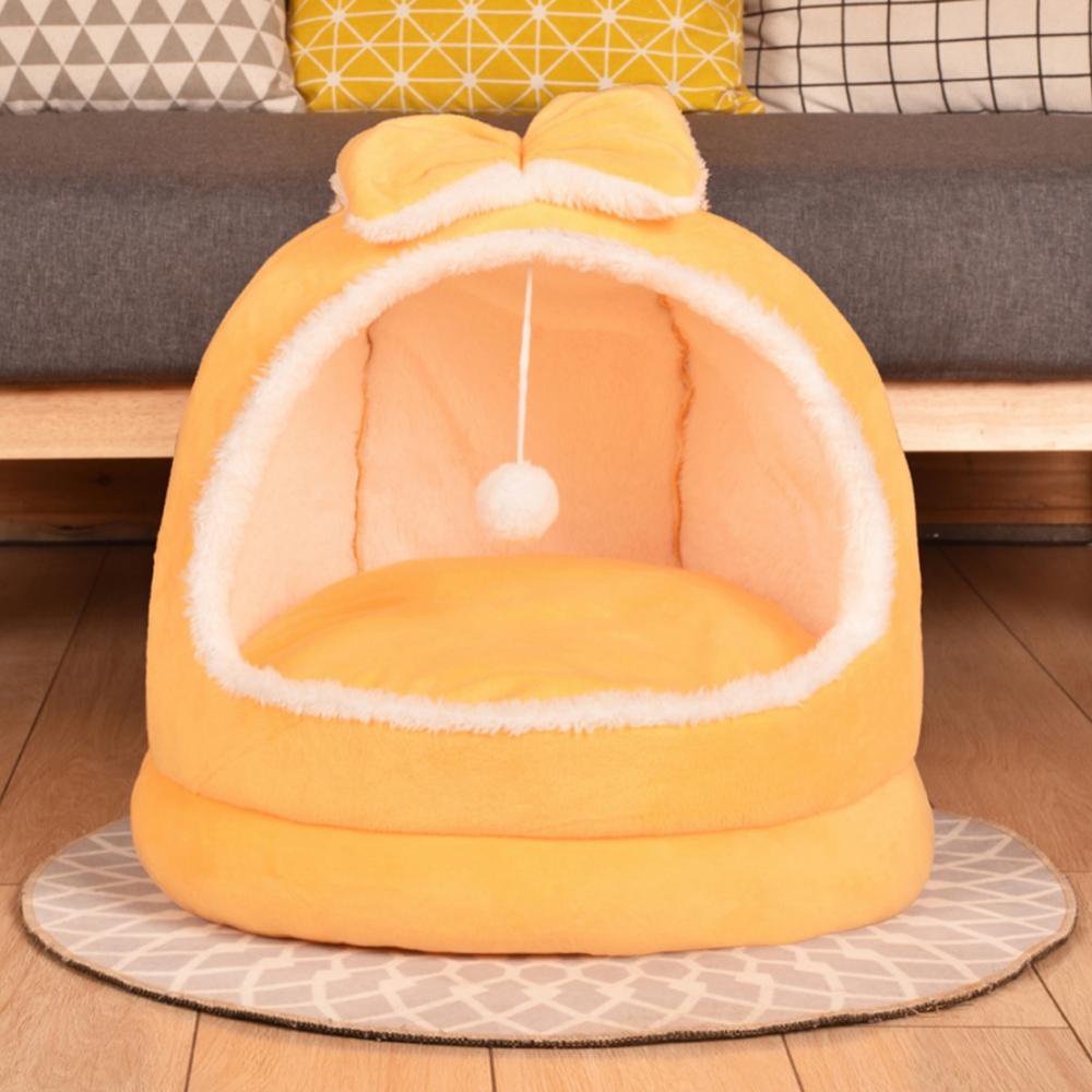 Yellow plush dome cat bed with matching bow on top, cream interior lining, and hanging white pom pom ball placed on a round grey mat in front of a grey sofa on hardwood floors