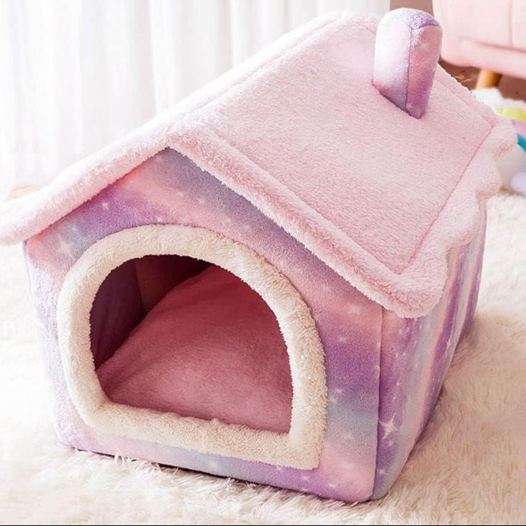A small house-shaped pet bed with a fluffy pink roof and trim, and pastel galaxy-print sides in pink, purple, and blue, shown on a soft white rug.