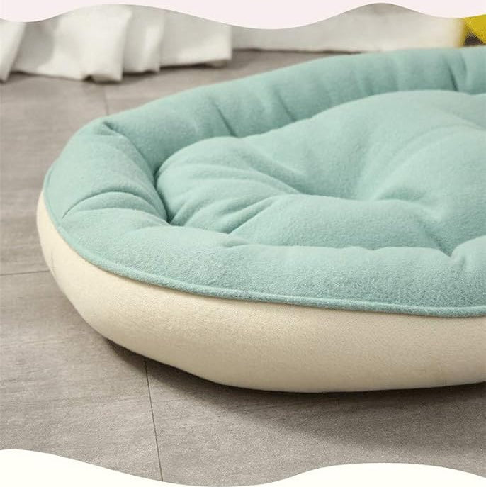 A close-up of a round pet bed with a light mint green cushioned top and a cream-colored base, placed on a gray tiled floor.