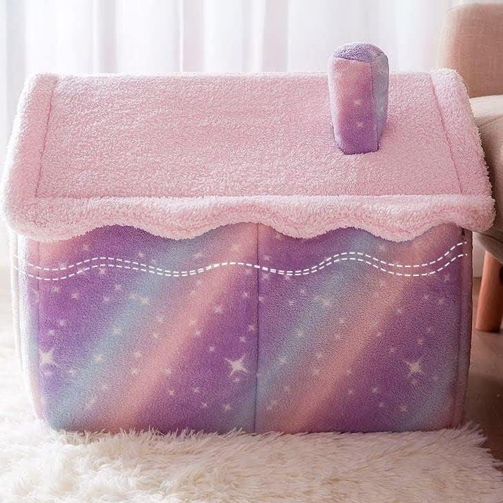 Pink and purple galaxy star print house-shaped pet bed with fluffy pink sherpa roof, scalloped trim, and chimney detail on a white fluffy rug in a soft pink room