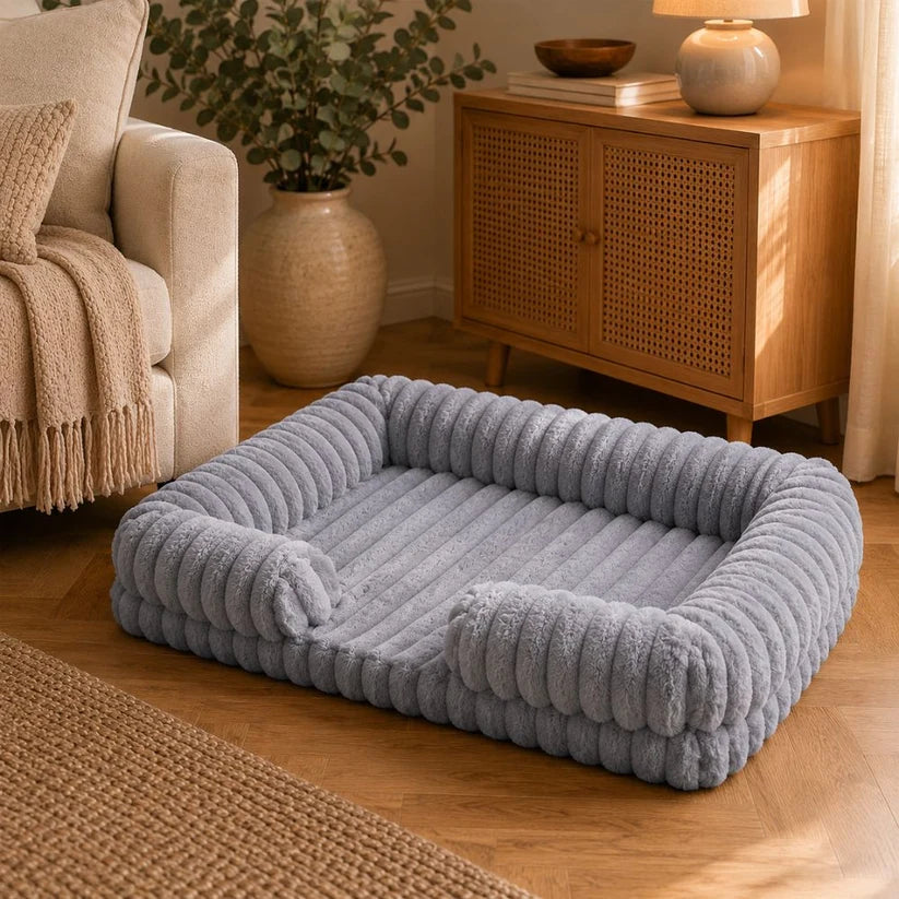 Dusty blue chunky corduroy pet bed on a herringbone wood floor in a warm, neutral living room styled with a cream sofa, rattan sideboard, and eucalyptus plant.
