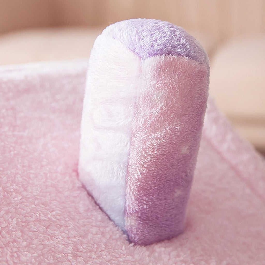 A close-up of a small plush cylindrical cushion or support pad in soft lavender and white, standing upright on a pink fuzzy blanket.