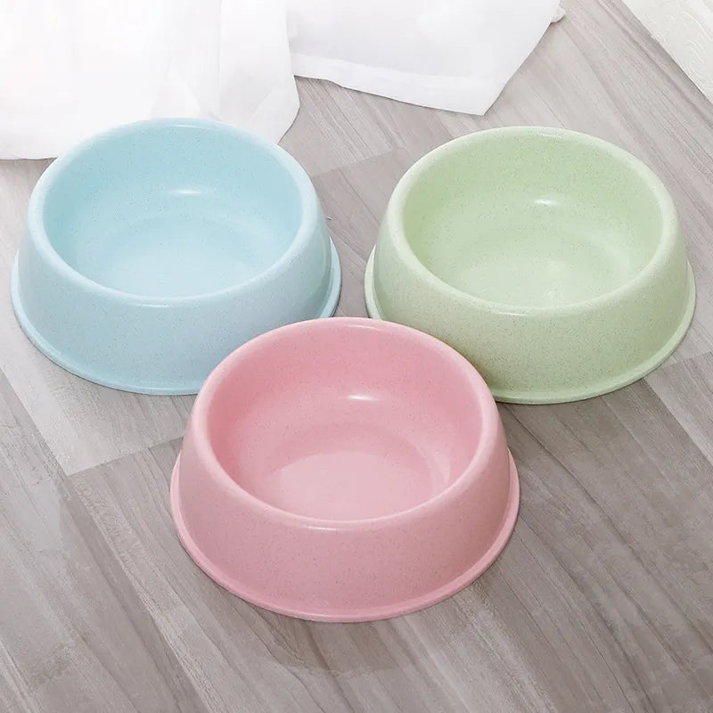 Three plastic pet bowls in pastel light blue, light green, and light pink, arranged on a gray wood-look floor with a white cloth in the background.