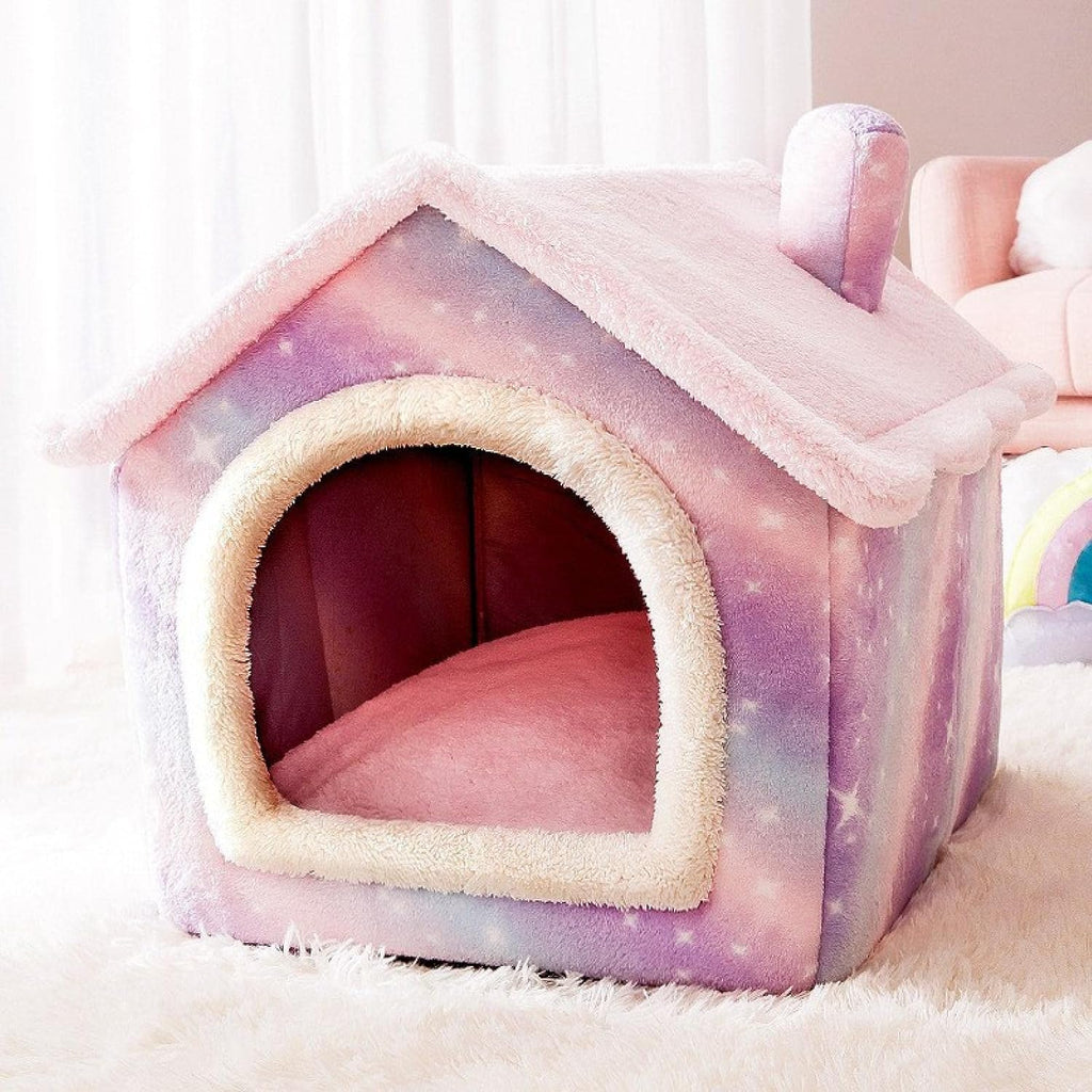 A small house-shaped pet bed with a pastel galaxy print in pink, purple, and blue, featuring a cream faux-fur trim around the arched entrance and a matching pink cushion inside, shown on a white rug in a softly lit room.