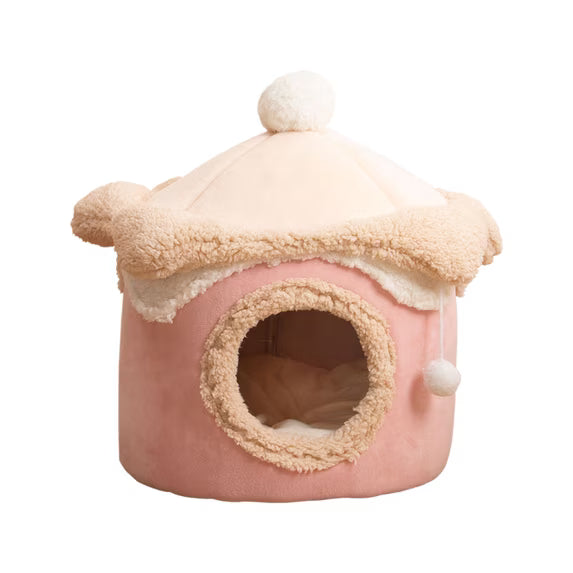 A pink and cream house-shaped pet bed with a plush dome roof, pom-pom details, and a circular opening on the front, shown on a plain white background.