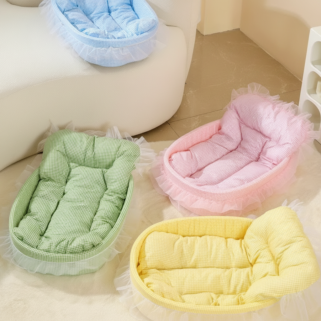 Four oval plush pet beds in pastel blue, pink, green, and yellow with gingham fabric and sheer lace ruffle trim displayed in a bright minimalist room scene