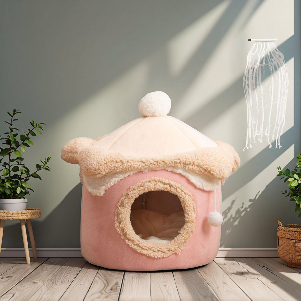 A pink plush pet house with a cream-colored fluffy roof and pom-pom details, set on a wooden floor against a gray wall with decorative plants and shadows.