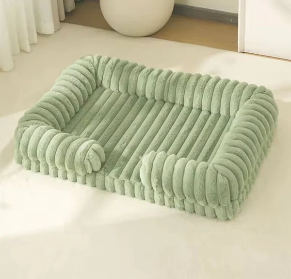 Sage green chunky corduroy pet bed on a cream floor in a minimal, light-filled room with a white sphere ornament in the background.