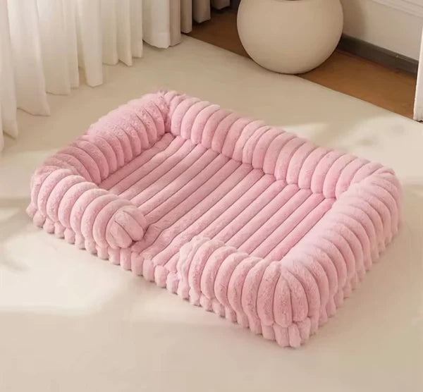 Blush pink chunky corduroy pet bed on a cream floor in a minimal, light-filled room with sheer curtains and a white sphere ornament in the background.