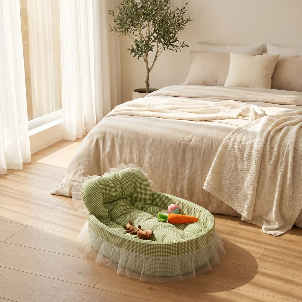 Sage green gingham oval pet bed with tufted bolster backrest, sheer white lace ruffle trim, and plush pet toys inside, displayed on a wood floor in a bright minimalist bedroom
