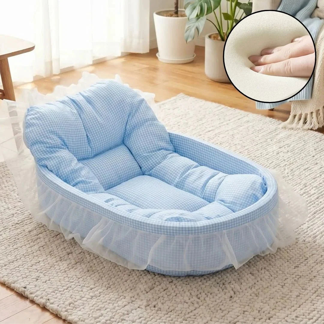 Pastel blue gingham oval pet bed with tufted bolster backrest and sheer white lace ruffle trim displayed on a cream rug in a bright living room, with inset close-up showing memory foam cushion material