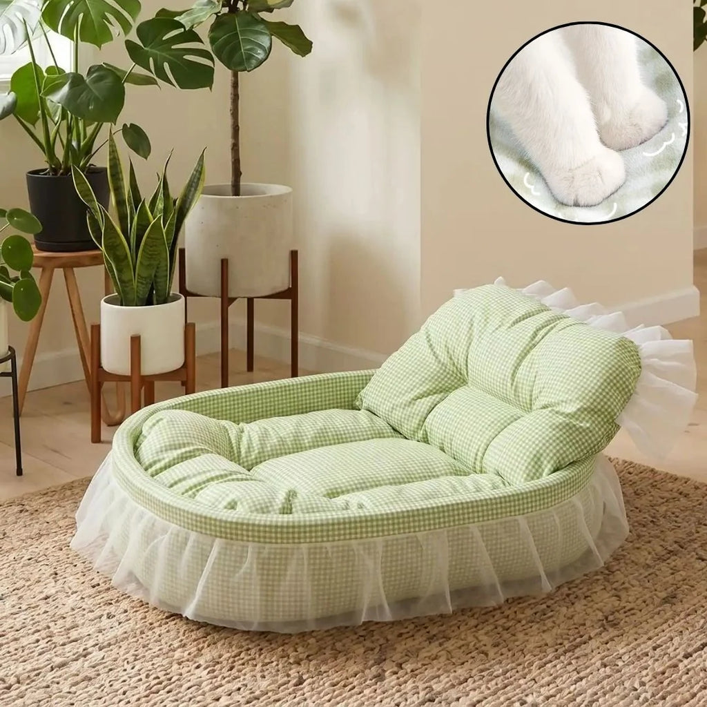 A light green and white gingham pet lounger with a white ruffled skirt, styled in a cozy room with potted plants. A circular inset in the top right shows a close-up of a white cat lying on the cushion.
