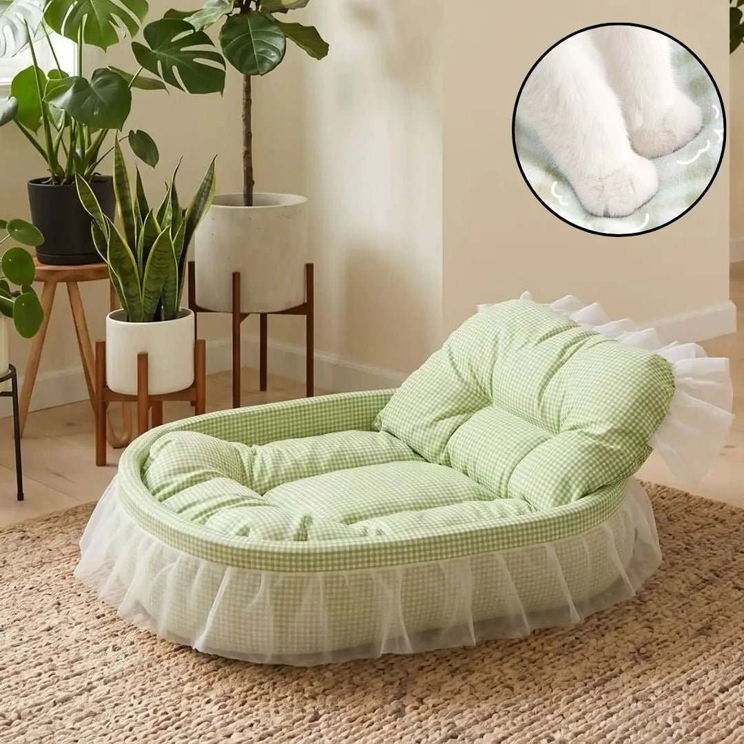 A light green and white gingham pet lounger with a white ruffled skirt, styled in a cozy room with potted plants. A circular inset in the top right shows a close-up of a white cat lying on the cushion.