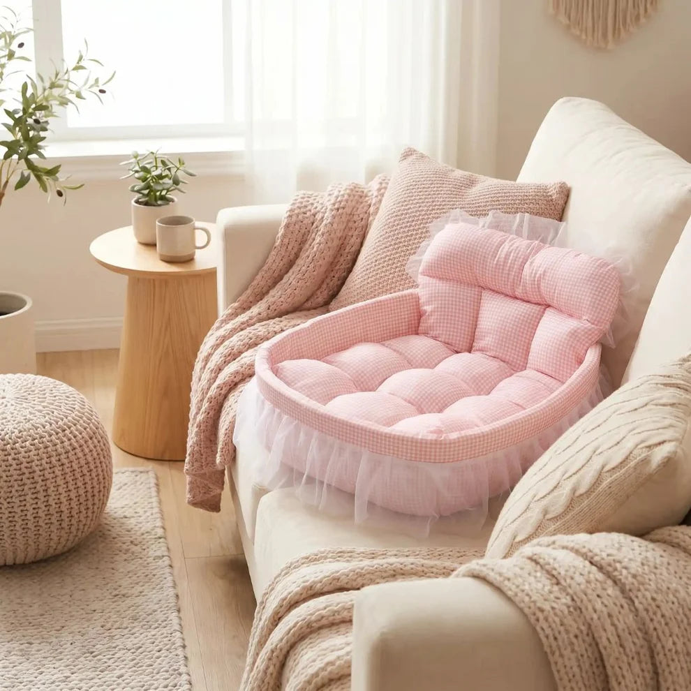 A pink tufted cushioned pet bed with a ruffled tulle skirt sits on a cream sofa in a softly lit living room. The bed includes a matching pink headrest cushion and is styled with knit blankets and pillows alongside a small wooden side table and window.