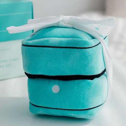 A turquoise velvet jewelry box dog toy with black piping and a white ribbon bow, photographed on a soft white surface with a blurred turquoise box in the background.