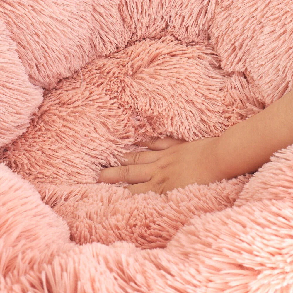 Close-up of a blush pink round donut pet bed with long shaggy faux fur, shown with a hand pressing into the plush center cushion to demonstrate softness