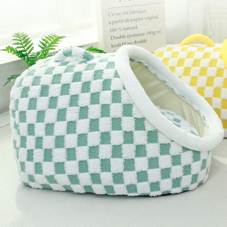 Sage green and white checkerboard plush pet cave bed with padded handbag-style carry handle, shown alongside yellow and white checkerboard cave bed in a bright lifestyle setting with greenery