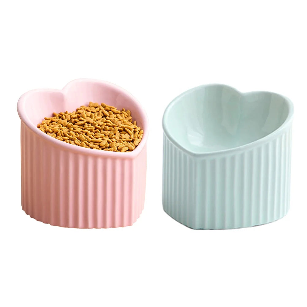Two ceramic heart-shaped pet bowls with ribbed exteriors in pink and mint green, pink bowl filled with dry kibble, white background

