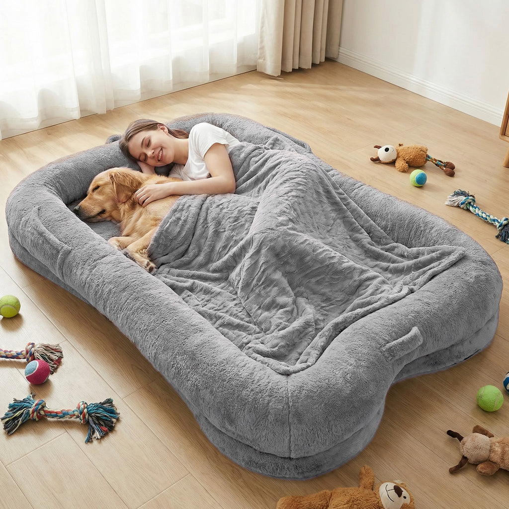 Woman and Golden Retriever cuddling together on an oversized grey plush human-sized dog bed with bone-shaped bolster sides and matching blanket surrounded by dog toys in a modern living room