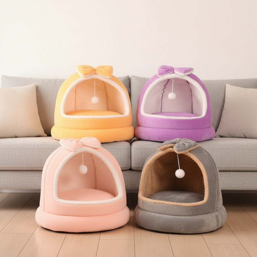 Four plush dome cat beds with bows and hanging white pom pom balls in yellow, purple, pink, and grey arranged in front of a grey sofa in a bright modern living room setting
