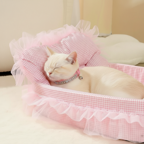 White cat wearing a pink crystal and bell collar sleeping in a pink gingham oval pet bed with fluffy pink feather trim and sheer white lace ruffle, cream background