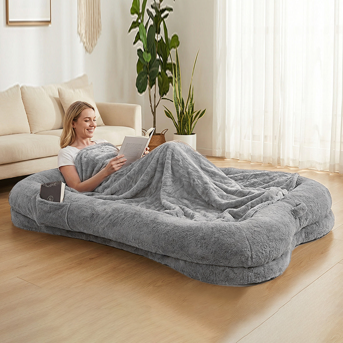Woman reading a book with blanket on an oversized grey plush human-sized dog bed with bone-shaped bolster sides and side pocket in a bright modern living room