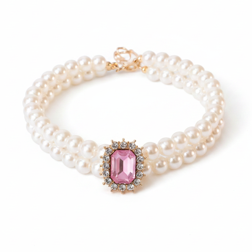 Double-strand white pearl pet collar with a large rectangular pink gemstone centerpiece surrounded by a crystal halo in gold setting, gold lobster clasp closure with a small crystal drop charm, laid in a circular shape on a white background — real product photo