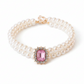 Double-strand white pearl pet collar with a large rectangular pink gemstone centerpiece surrounded by a crystal halo in gold setting, gold lobster clasp closure with a small crystal drop charm, laid in a circular shape on a white background — real product photo