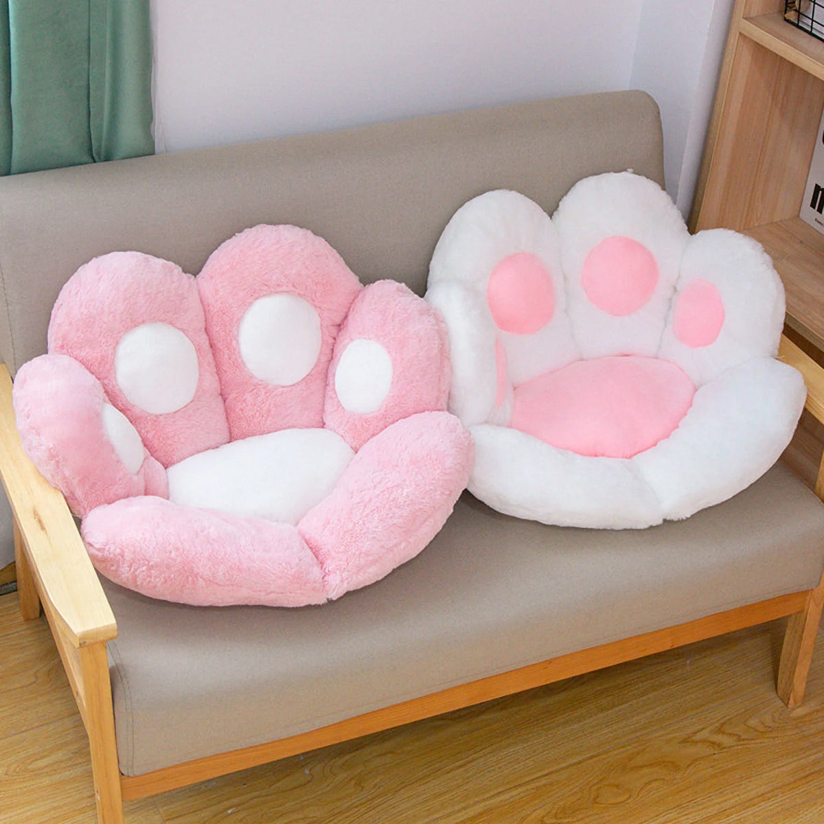 Lifestyle image showing two plush paw-shaped pet bed cushions side by side on a taupe linen sofa with a light oak wood frame — one blush pink with white toe pads and one white with pink toe pads — in a bright casual living room with teal curtains and a wooden bookshelf