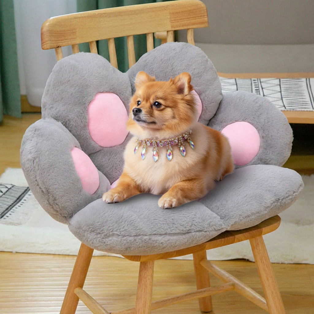 Golden Pomeranian wearing a silver and rainbow crystal marquise drop collar sitting inside a grey and pink plush paw-shaped pet bed cushion placed on a natural wood spindle chair, with a cream geometric area rug, teal curtains, and a light grey wall in the background