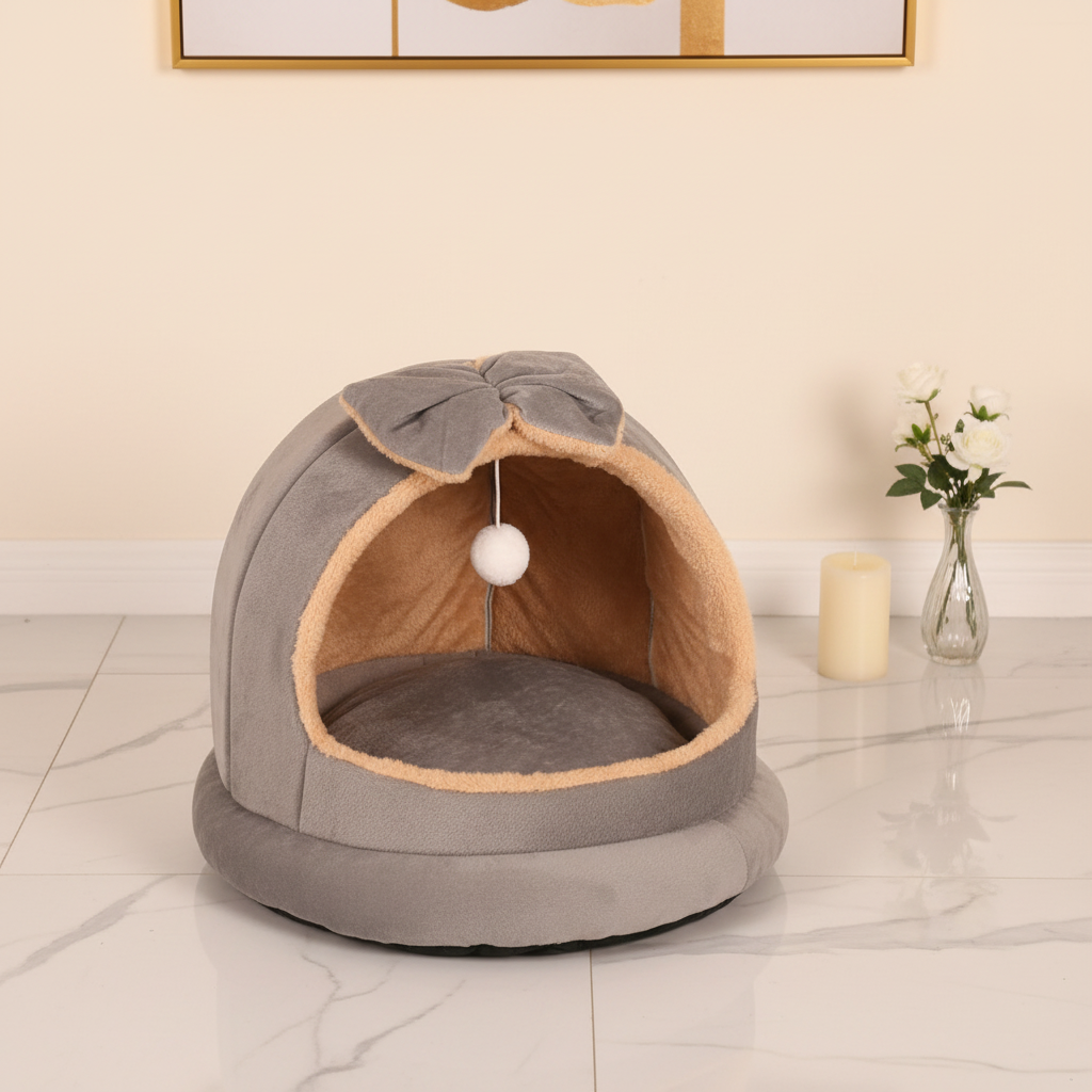 Grey suede plush dome cat bed with bow on top and hanging white pom pom ball displayed in an elegant interior setting with white marble floors, cream walls, gold-framed art, pillar candle, and white floral arrangement