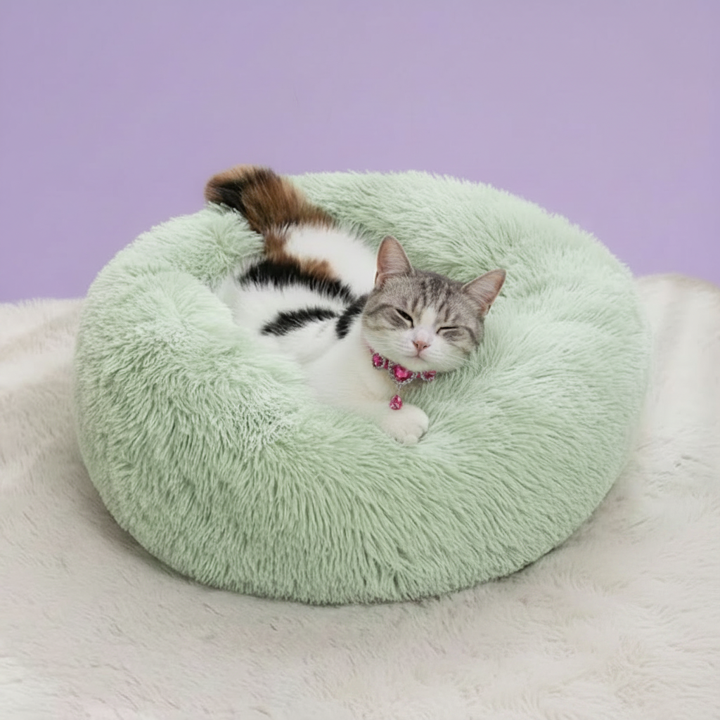 Grey and white tabby cat wearing a pink rhinestone collar napping contentedly in a sage green round donut pet bed with long shaggy faux fur, set against a soft lavender backdrop