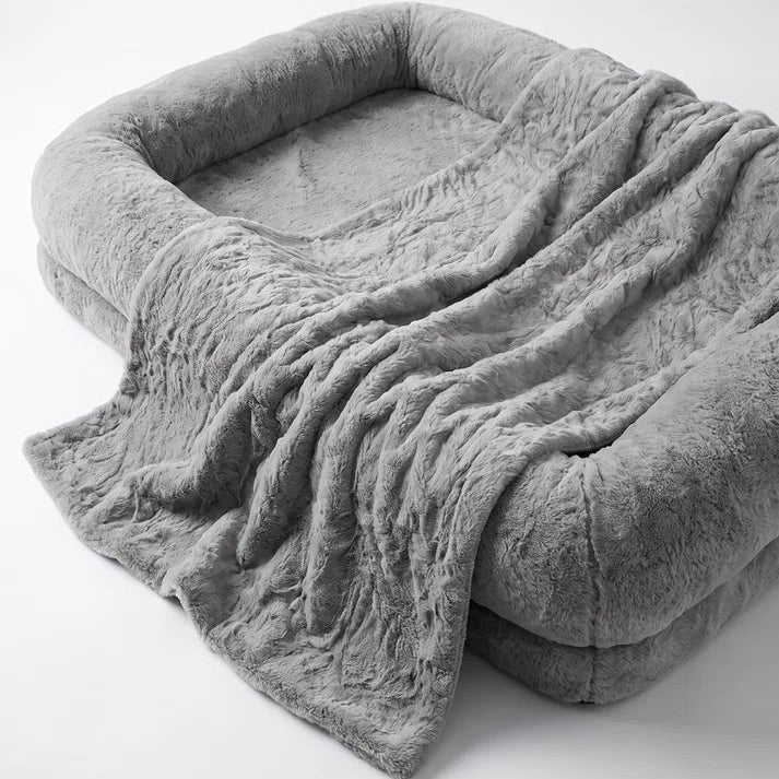 Close-up of oversized grey plush human-sized dog bed with bolster sides and draped faux fur blanket showing texture detail on white background