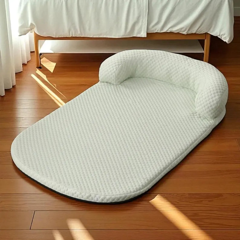 A light sage green textured pet bed cushion with a built-in headrest, placed on a wooden floor next to a bed with white bedding.