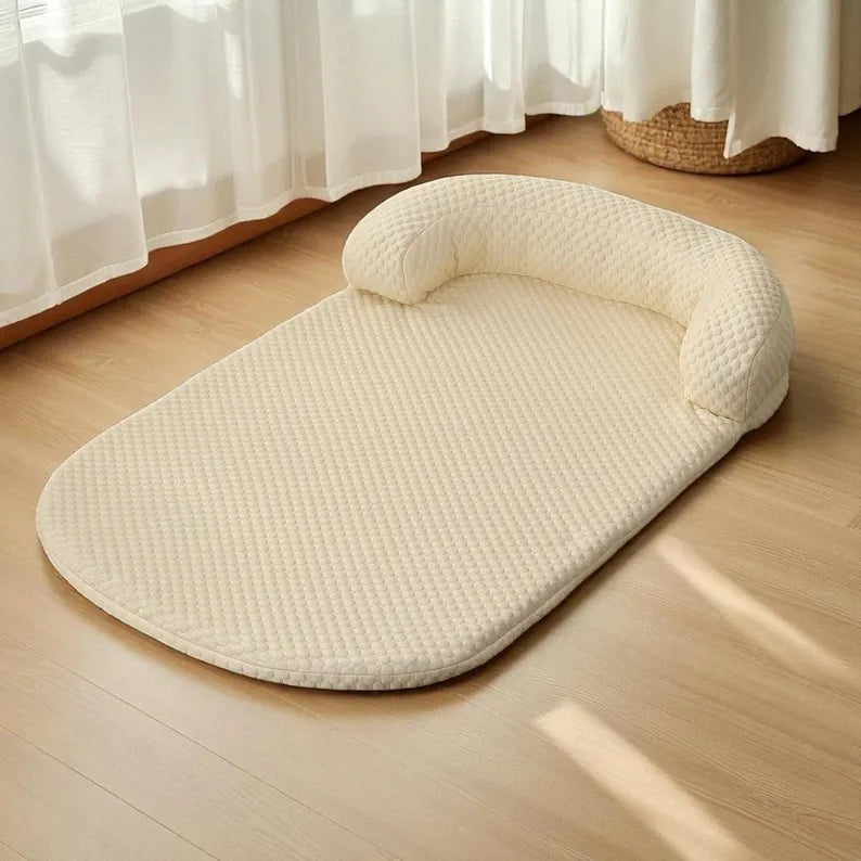 A cream-colored textured pet bed mat with a built-in curved headrest, placed on a light wood floor near white curtains.