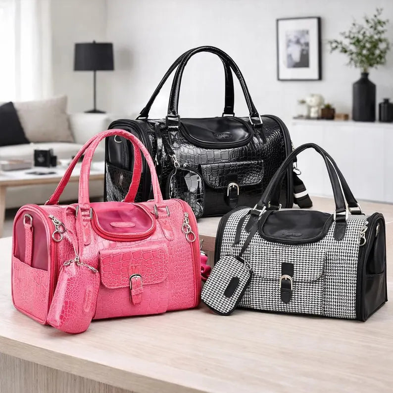 Three designer-style pet carrier displayed on a light wood table in a modern living room setting: a bright pink crocodile-embossed tote bag with a matching pouch, a glossy black crocodile-embossed tote bag, and a black-and-white houndstooth pet carrier bag with a matching pouch.