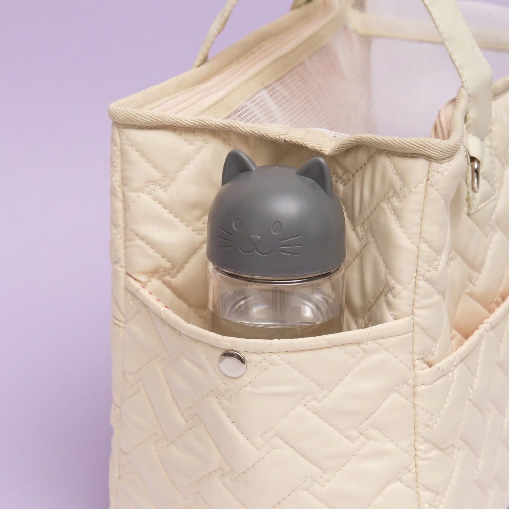 A close-up of a quilted cream tote bag with a front pocket holding a clear bottle with a gray cat-shaped lid, against a soft lavender background.