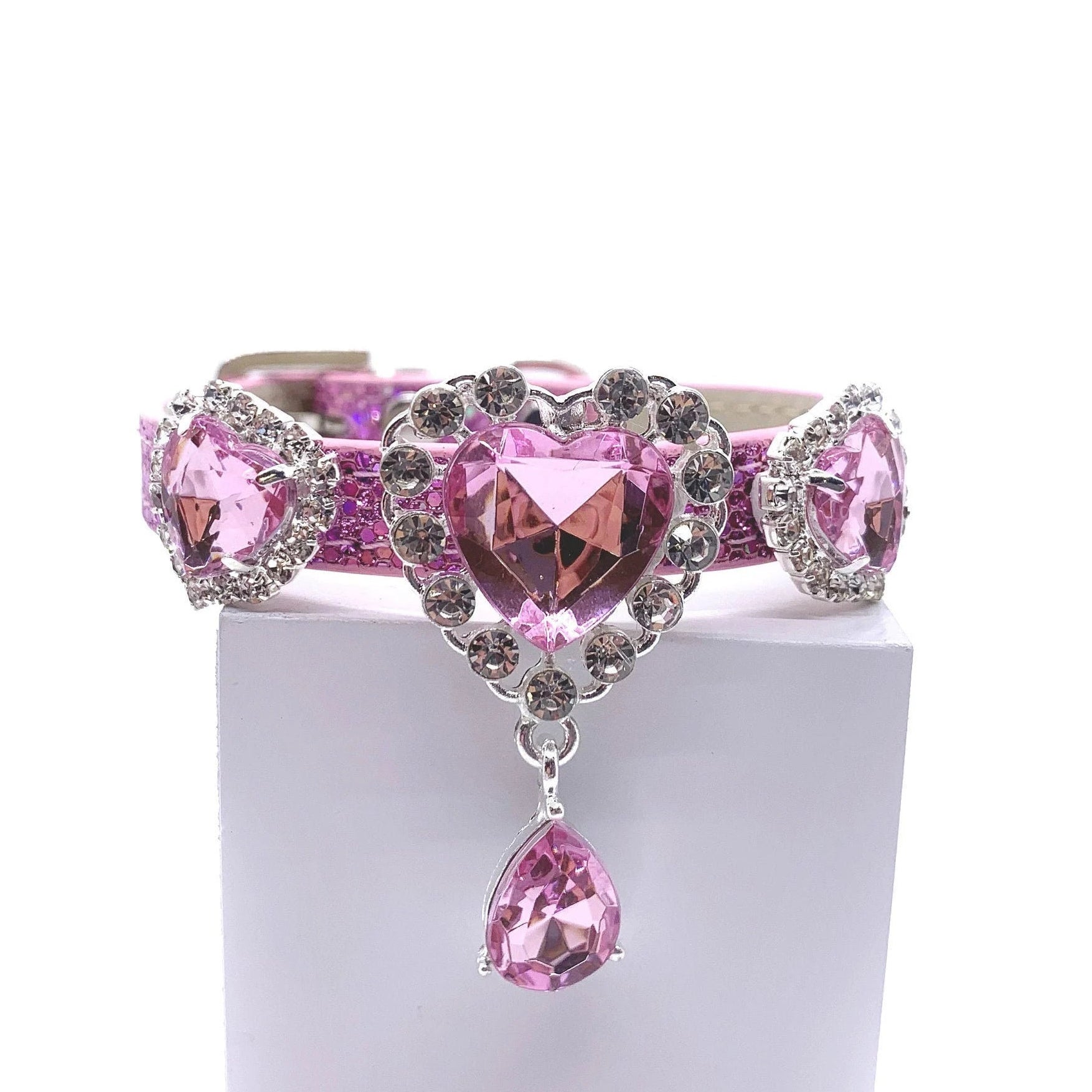 Pink glitter pet collar with three rose pink heart crystal charms in rhinestone halo settings, center charm with dangling pink teardrop crystal drop, laid flat showing full length with rhinestone-encrusted buckle for cats and dogs