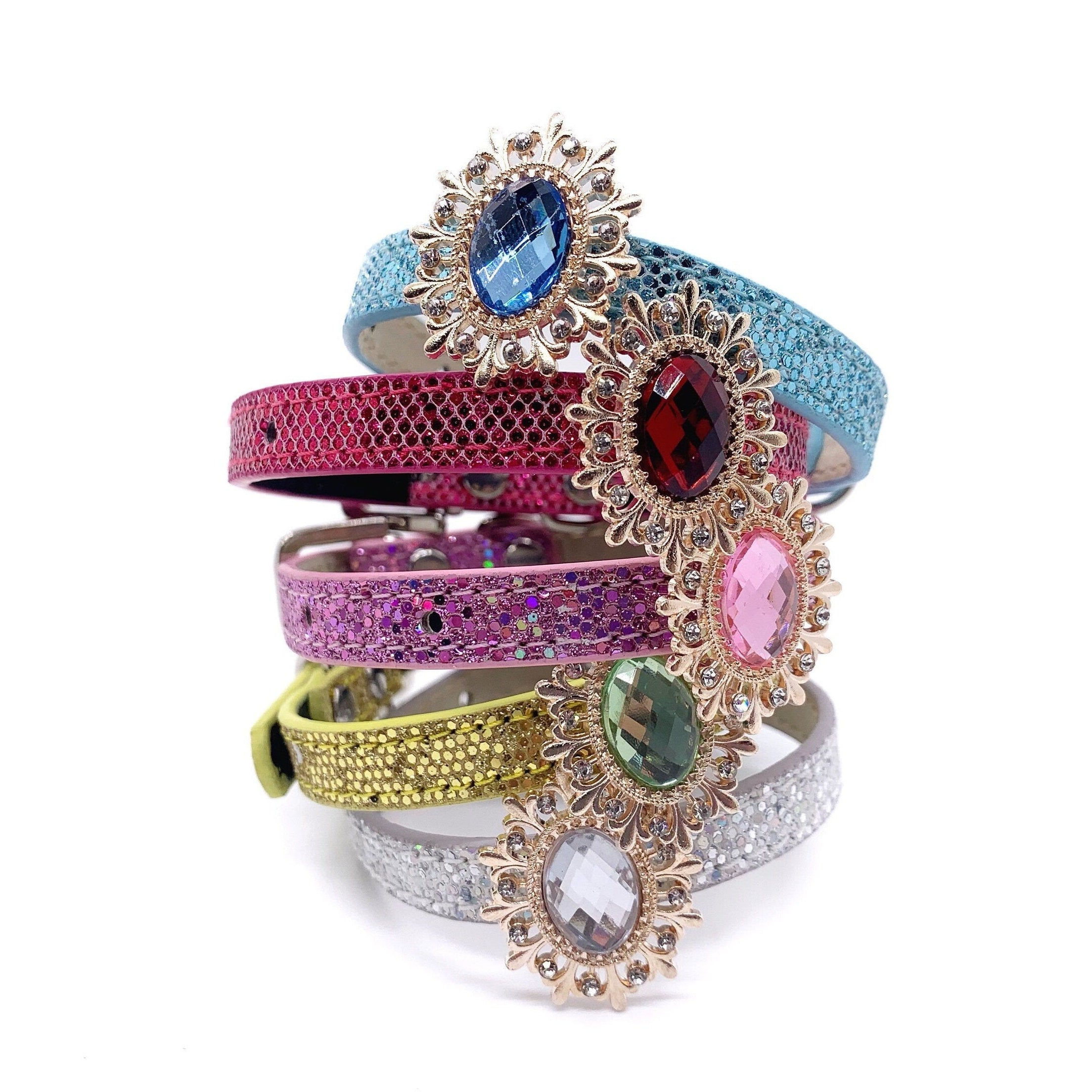 Stacked luxury pet collar collection featuring baby blue with sapphire oval brooch, hot pink honeycomb with ruby oval brooch, lavender glitter with rose pink oval brooch, yellow citrine with peridot green oval brooch, and silver glitter with clear crystal oval brooch, all with gold ornate sunburst frames for cats and dogs