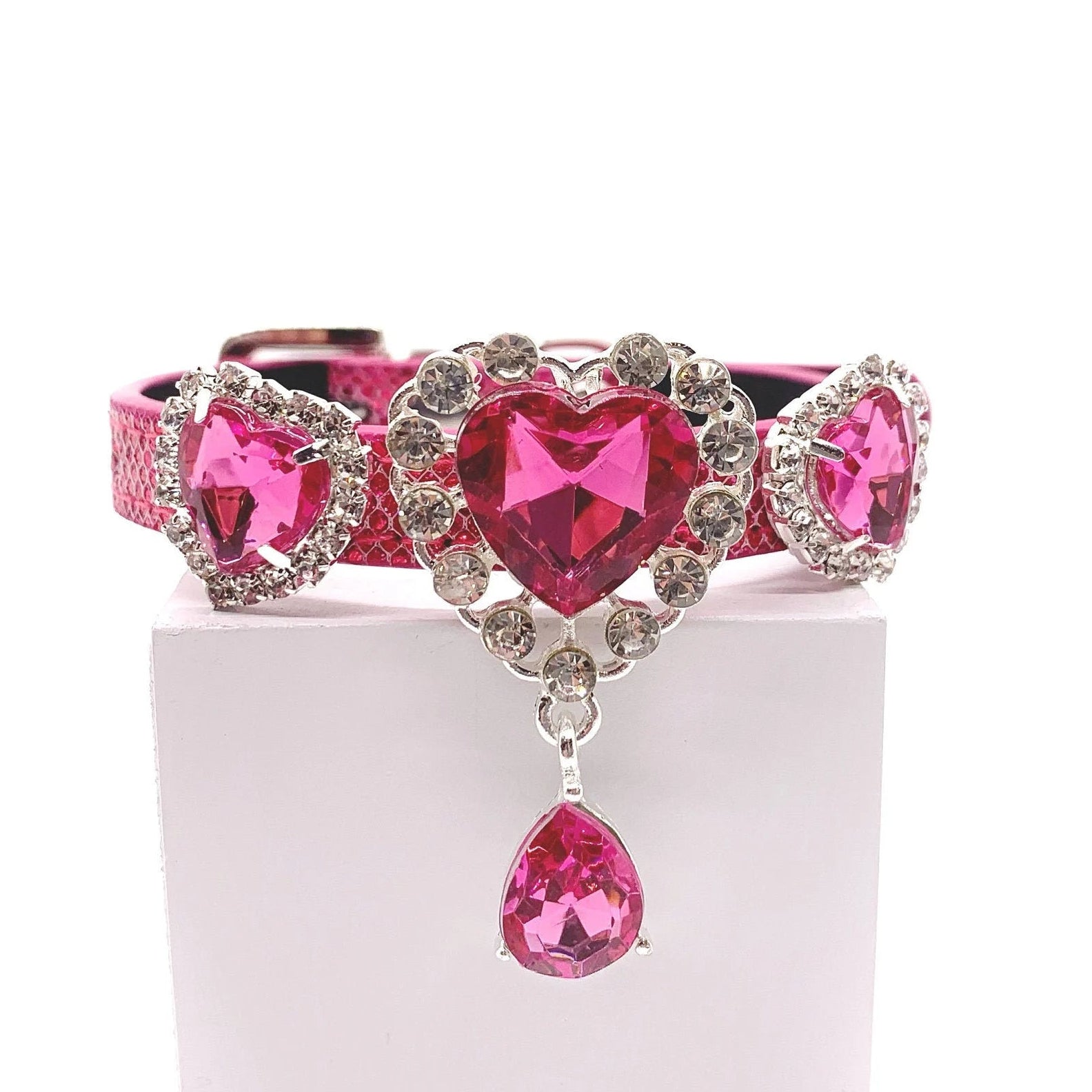 Hot pink metallic glitter pet collar with three fuchsia heart crystal charms in rhinestone halo settings, center charm with dangling pink teardrop crystal drop, displayed on stand for cats and dogs