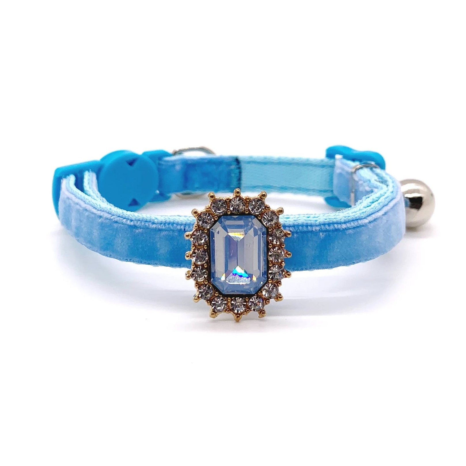 Baby blue velvet pet collar with light blue emerald-cut rhinestone charm, gold halo setting, and silver bell on white background