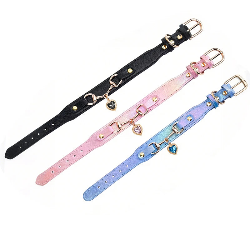 Three pet collars in black, iridescent pink, and holographic blue-purple with gold buckle hardware and crystal heart charm pendants, displayed on a white background