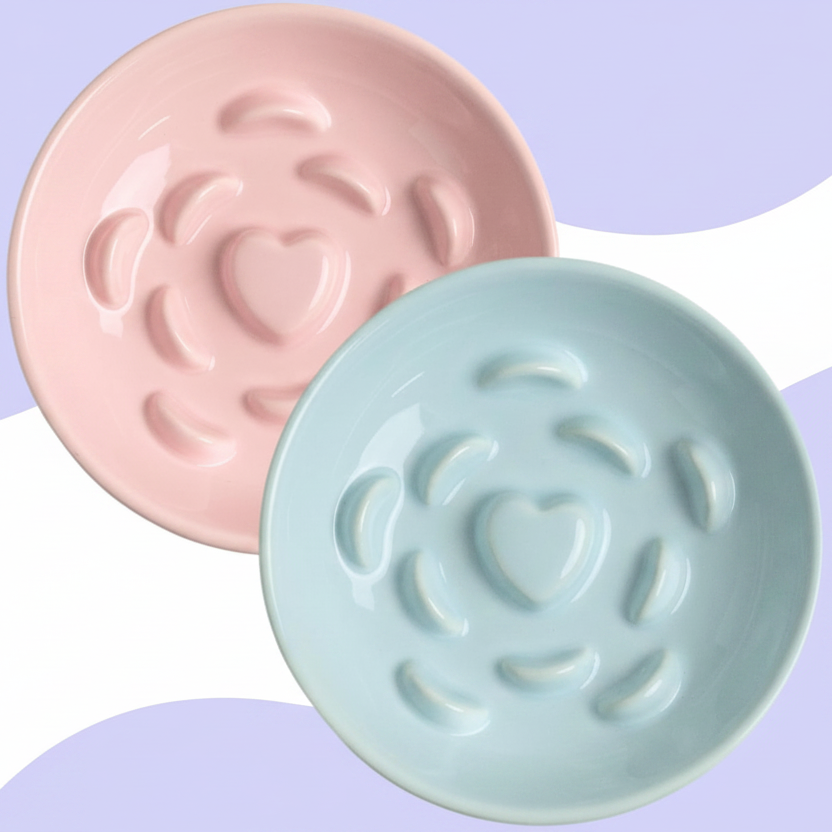 Pink and mint ceramic raised slow feeder cat bowls with heart-center ridges on a soft lavender wave background