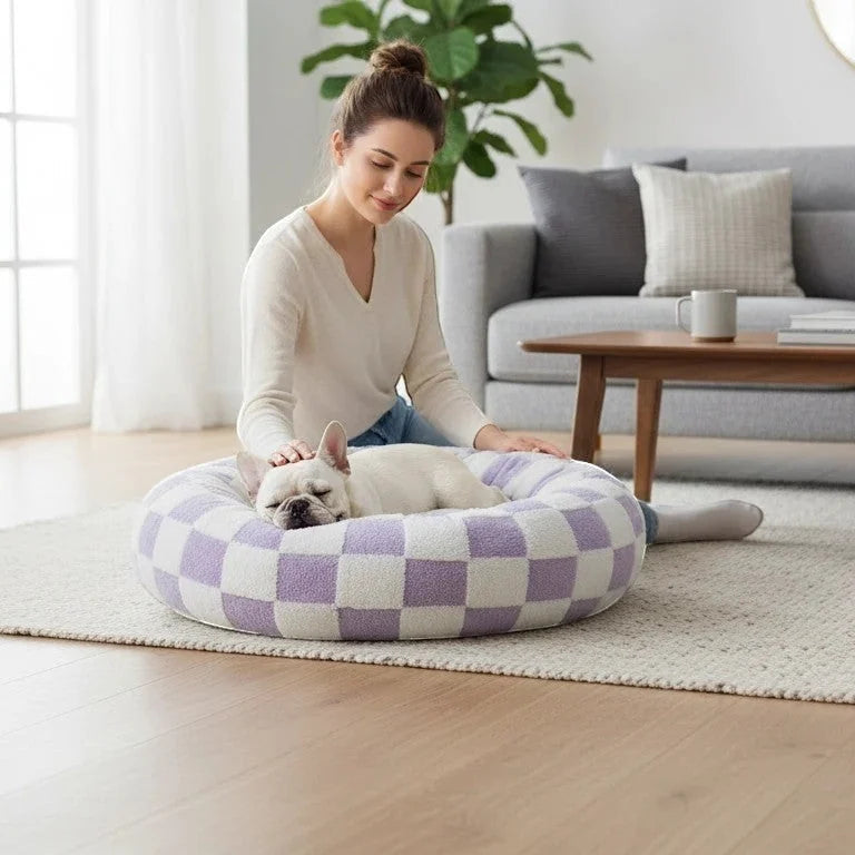 Lifestyle image of a smiling brunette woman in a cream sweater and jeans sitting cross-legged on a cream area rug, petting a sleeping white French Bulldog curled inside a large lavender and cream checkered plush round donut pet bed, in a bright modern living room with a grey sofa, walnut coffee table, fiddle leaf fig plant, and sheer white curtains