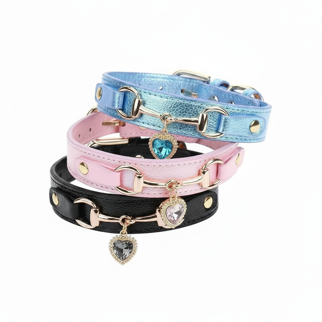 Three stacked pet collars in metallic blue, pink, and black leather with gold snaffle-bit hardware and crystal heart charm pendants in blue, clear, and grey, displayed on a white background