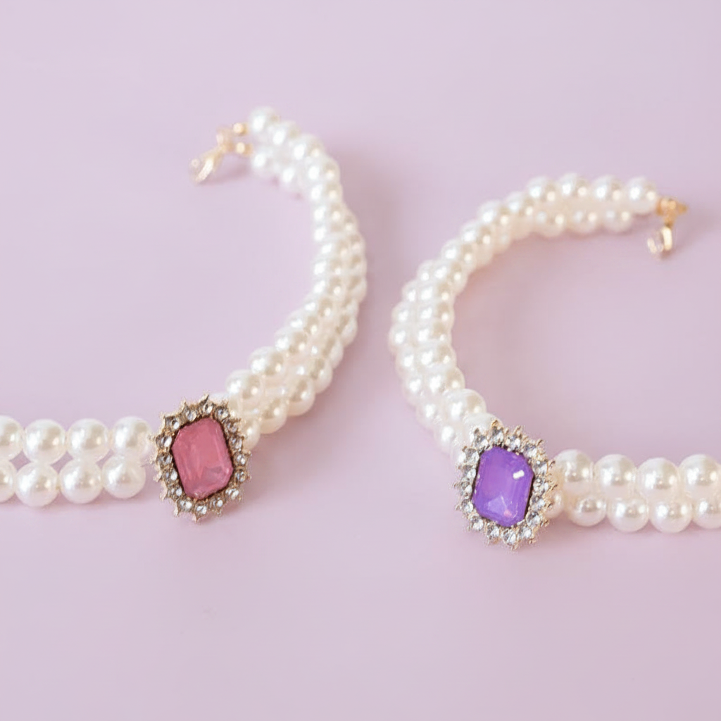 Two double-strand white pearl pet collars laid flat side by side — one with a pink oval gemstone centerpiece surrounded by a crystal halo, and one with a purple/lavender oval gemstone centerpiece surrounded by a crystal halo — both with silver hardware, on a soft lavender background — real product photo